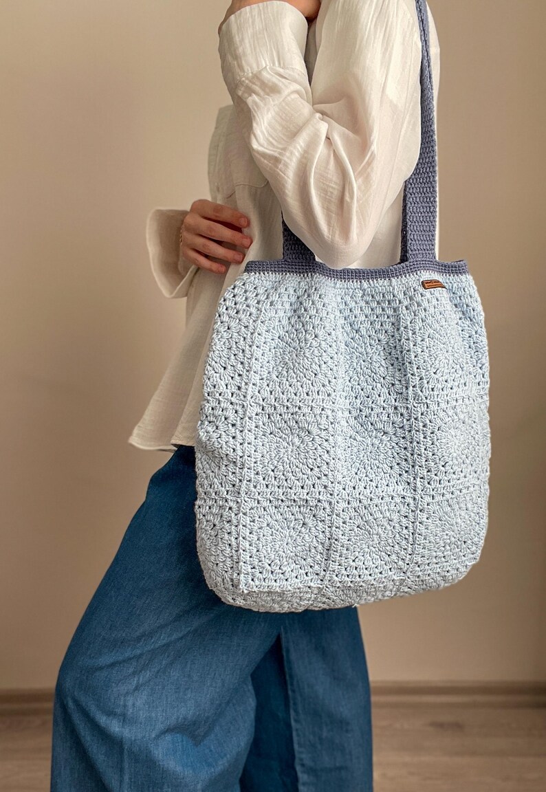 Puede incluir: Un bolso tote de crochet azul claro con una correa azul oscuro. El bolso est&aacute; hecho con un patr&oacute;n de puntadas texturizadas.
