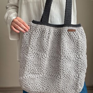 Puede incluir: Una bolsa de mano de crochet gris con correas negras. La bolsa tiene una etiqueta de cuero marr&oacute;n con el texto "Handmade with Love" en ella.