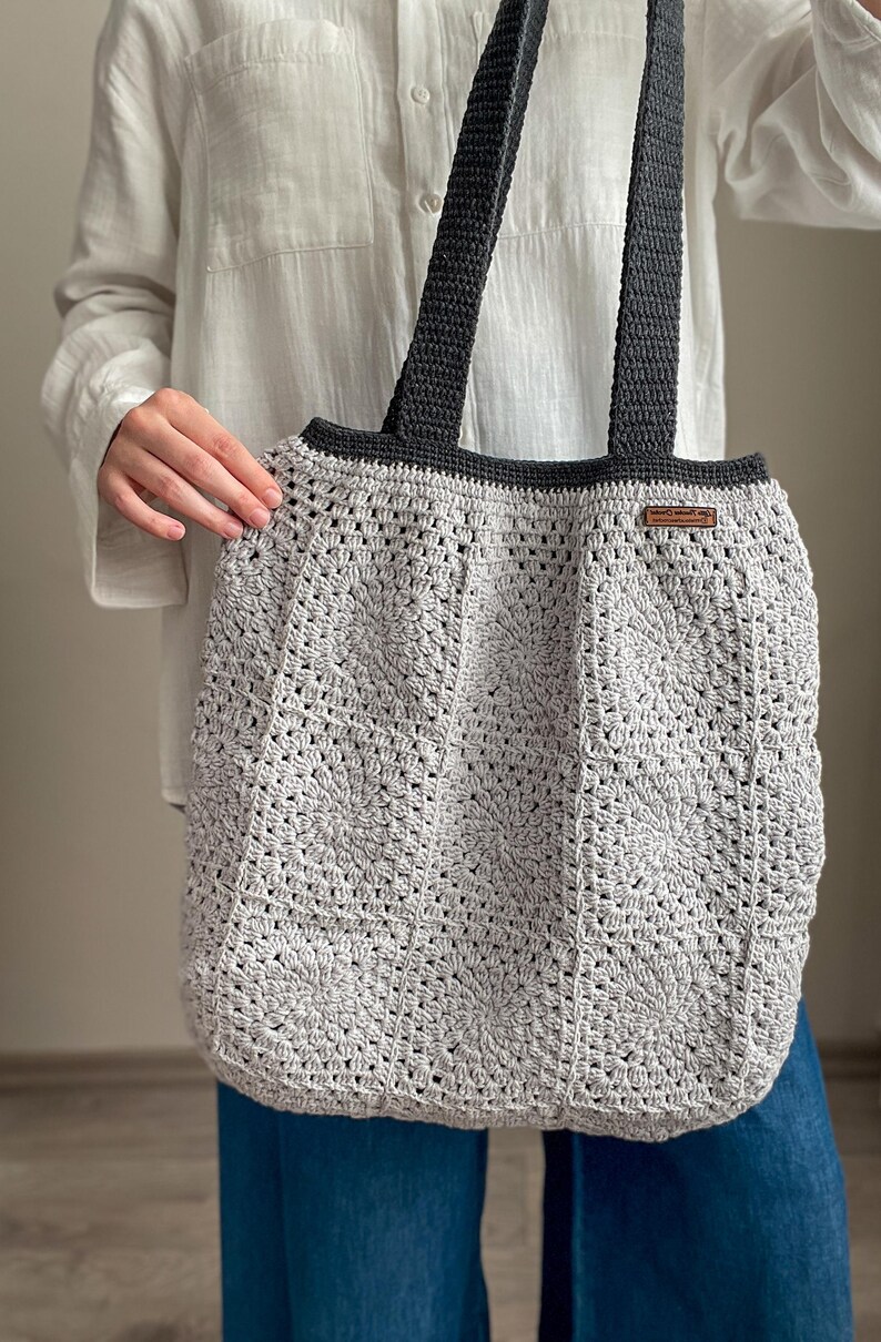 Puede incluir: Una bolsa de mano de crochet gris con correas negras. La bolsa tiene una etiqueta de cuero marr&oacute;n con el texto "Handmade with Love" en ella.