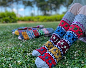 May include: A pair of colorful knitted socks with geometric patterns in red, blue, yellow, and white. The socks are on a grassy surface with other socks in the background. The socks have gray toes and cuffs.