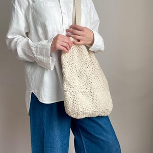 Puede incluir: Una bolsa de mano de crochet blanca con una correa gruesa. La bolsa la lleva una persona que lleva una camisa blanca abotonada y pantalones vaqueros azules.