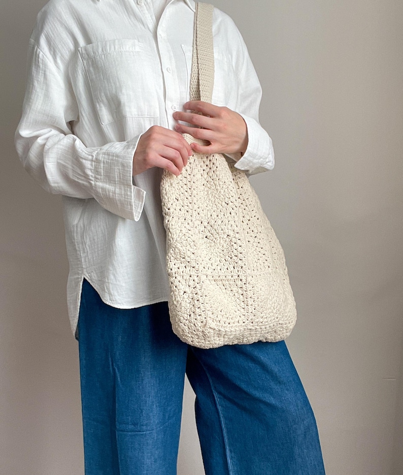 Puede incluir: Una bolsa de mano de crochet blanca con una correa gruesa. La bolsa la lleva una persona que lleva una camisa blanca abotonada y pantalones vaqueros azules.