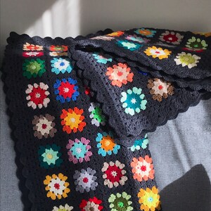 May include: A colorful granny square crochet blanket with a dark gray background. The blanket is made up of many different colored squares, including red, orange, yellow, green, blue, and purple.