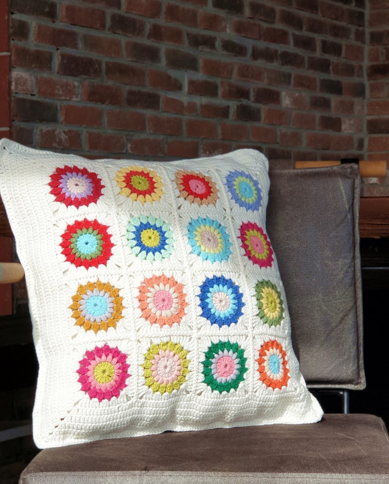 K&ouml;nnte beinhalten: Ein wei&szlig;es H&auml;kelkissen mit einem bunten geometrischen Muster. Das Kissen besteht aus 16 Quadraten, jedes mit einem andersfarbigen Blumendesign. Das Kissen liegt auf einem braunen Stuhl.