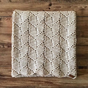 May include: A beige crocheted blanket with a diamond pattern. The blanket has a scalloped edge and a small brown leather tag.