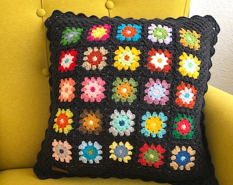 Könnte beinhalten: Ein schwarzes Häkelkissen mit einem gewellten Rand und einem bunten Blumenmuster. Das Kissen besteht aus mehreren Quadraten mit verschiedenen farbigen Blumen in jedem Quadrat.
