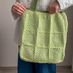 Puede incluir: Una bolsa de mano de crochet de color verde claro con una correa a juego. La bolsa est&aacute; hecha de cuadrados con un patr&oacute;n circular en el centro de cada cuadrado.