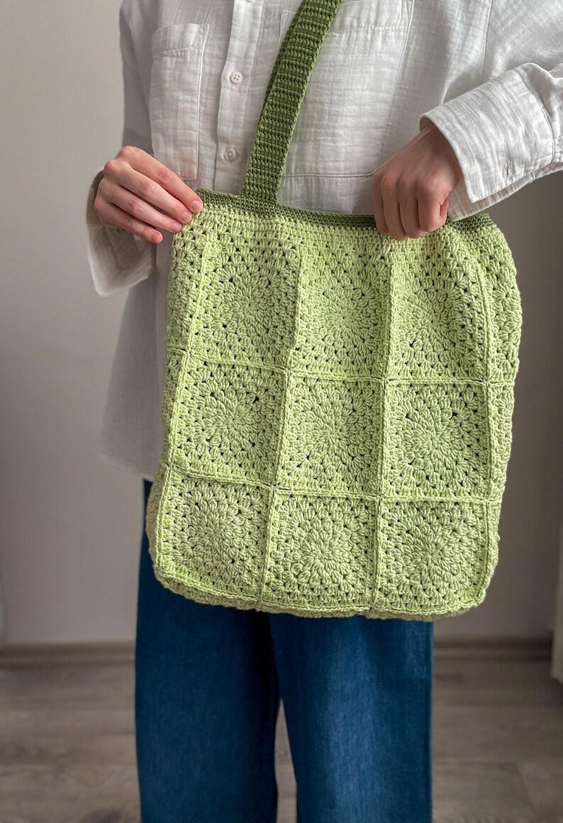 Puede incluir: Una bolsa de mano de crochet de color verde claro con una correa a juego. La bolsa est&aacute; hecha de cuadrados con un patr&oacute;n circular en el centro de cada cuadrado.