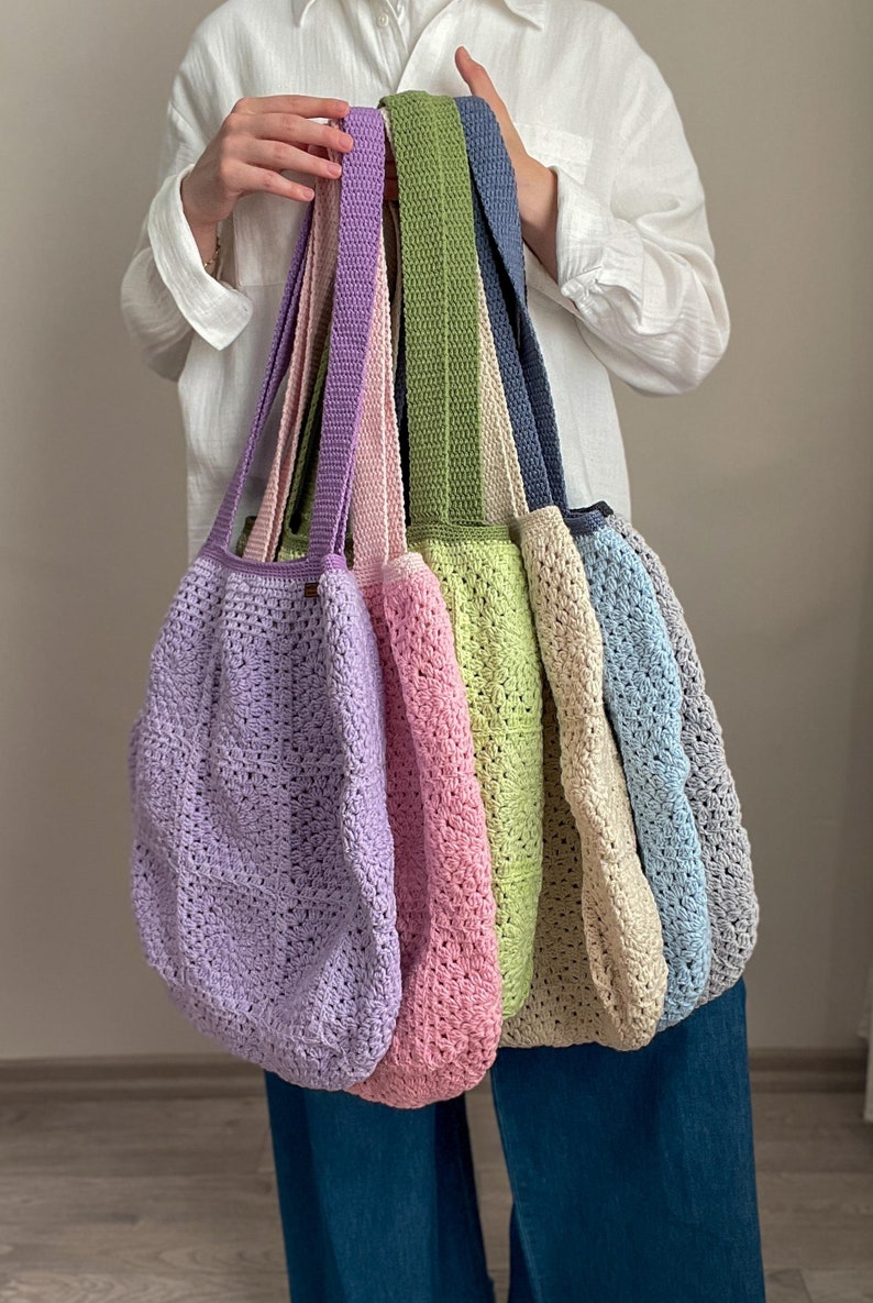 Puede incluir: Un conjunto de seis bolsas de mano de crochet en varios colores pastel, incluyendo lavanda, rosa, verde claro, beige y azul. Las bolsas tienen asas largas y est&aacute;n hechas con un delicado patr&oacute;n de tejido abierto.
