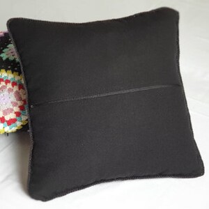 May include: A black pillow with a zipper closure. The pillow is lying on a white surface. A colorful crocheted pillow is partially visible behind the black pillow.