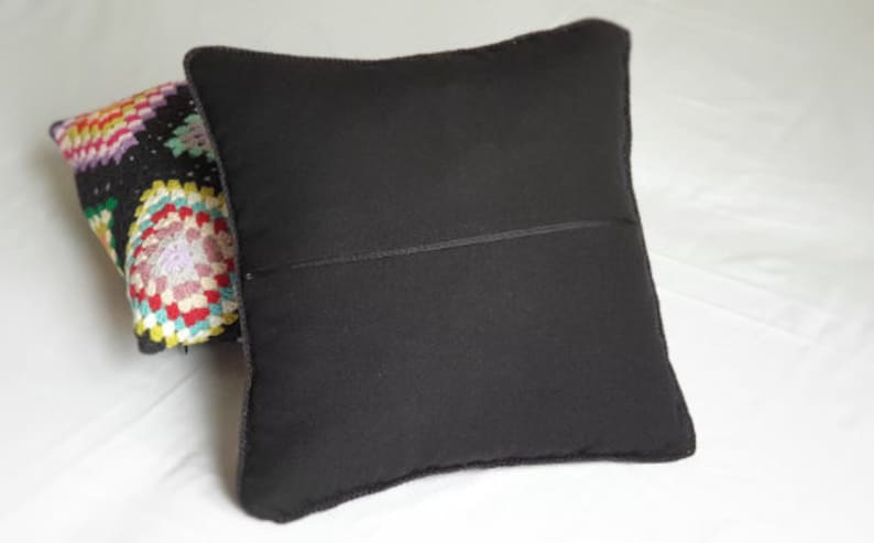 May include: A black pillow with a zipper closure. The pillow is lying on a white surface. A colorful crocheted pillow is partially visible behind the black pillow.