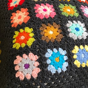 May include: A black crochet pillow with a colorful floral pattern. The pillow is made of multiple granny squares with different colored flowers. The pillow has a label that says "Little Touches Crochet" and "littletouchescrochet.com".