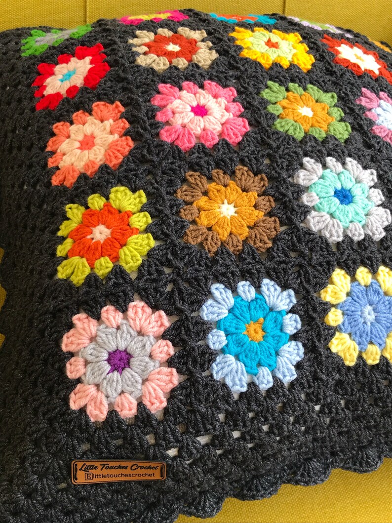 May include: A black crochet pillow with a colorful floral pattern. The pillow is made of multiple granny squares with different colored flowers. The pillow has a label that says "Little Touches Crochet" and "littletouchescrochet.com".