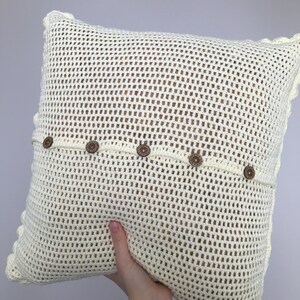 May include: A white crocheted pillow with a decorative border and brown buttons. The pillow is made of a mesh-like fabric.