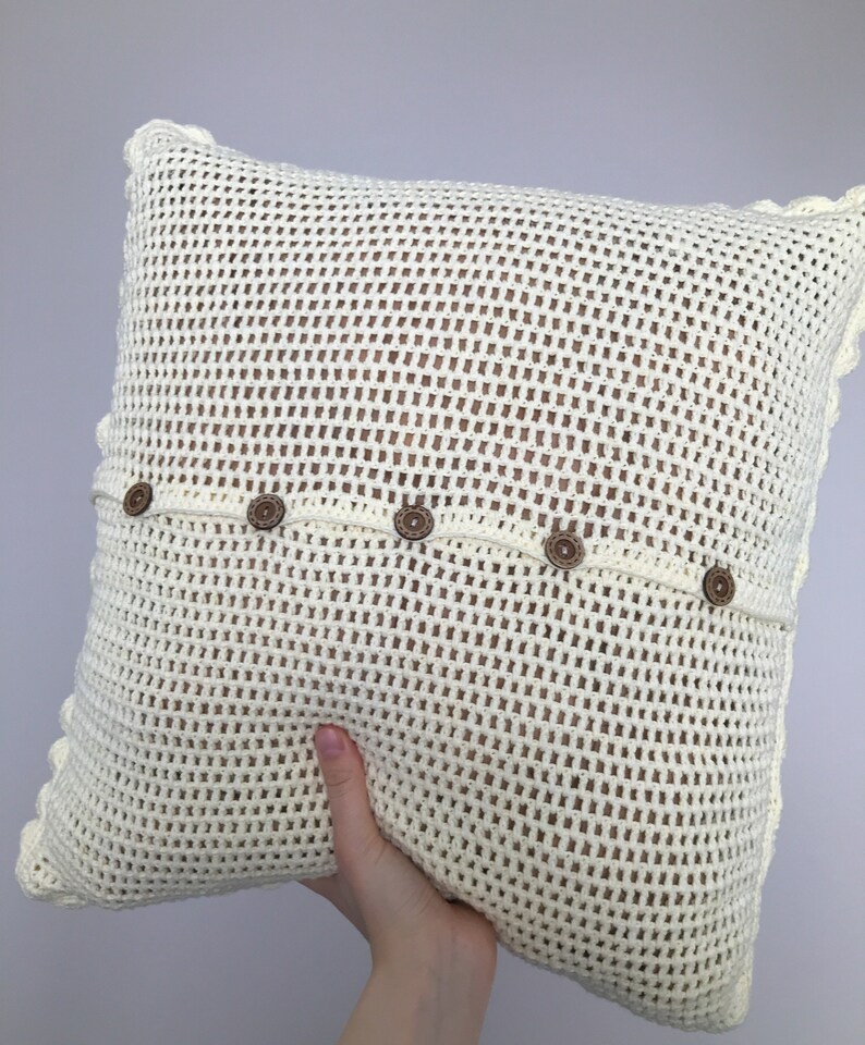 May include: A white crocheted pillow with a decorative border and brown buttons. The pillow is made of a mesh-like fabric.
