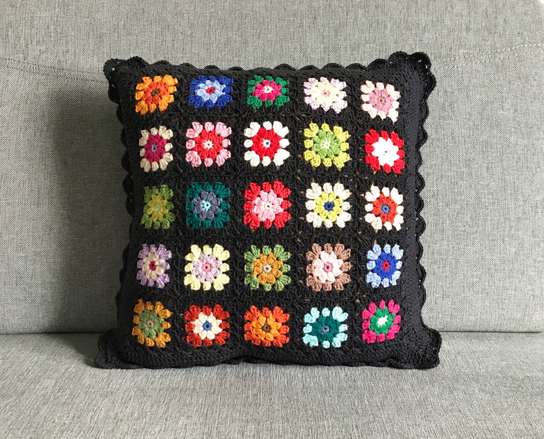 May include: A black crocheted pillow with a square pattern of colorful flowers. The pillow has a scalloped edge.