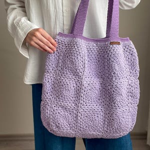 Puede incluir: Una bolsa de mano de crochet de color lavanda con una etiqueta de cuero marr&oacute;n. La bolsa tiene una parte superior ancha y abierta y dos asas largas.