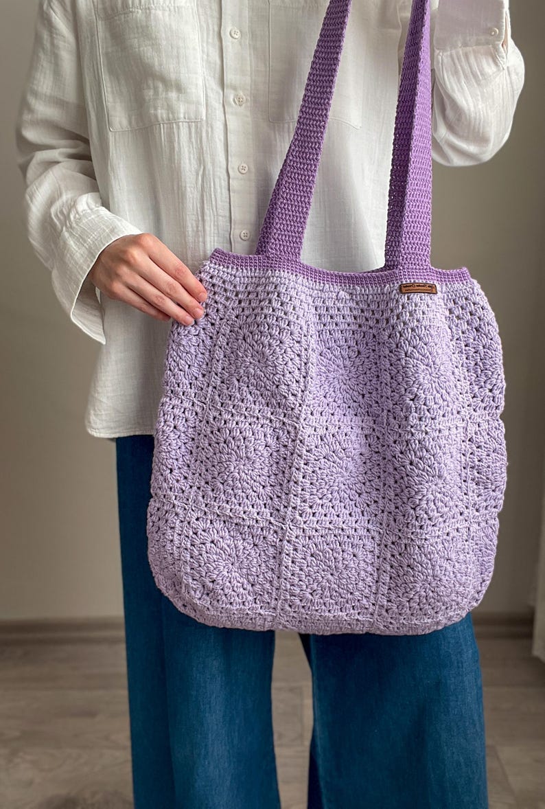 Puede incluir: Una bolsa de mano de crochet de color lavanda con una etiqueta de cuero marr&oacute;n. La bolsa tiene una parte superior ancha y abierta y dos asas largas.