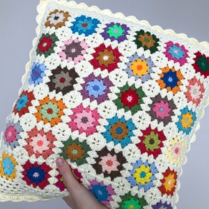 May include: A colorful crocheted pillow with a white background and a scalloped edge. The pillow is made up of many small squares, each with a different colored flower design.