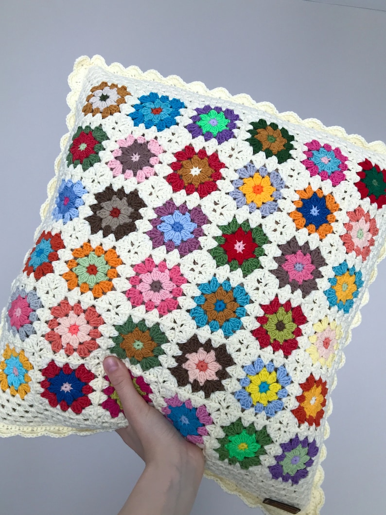May include: A colorful crocheted pillow with a white background and a scalloped edge. The pillow is made up of many small squares, each with a different colored flower design.