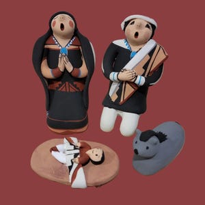 May include: Hand-painted ceramic figurines depicting a nativity scene. The set includes figures of a woman, a man, a baby, and a donkey. The figures are in various colors, including black, brown, white, and blue.