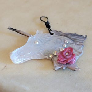 May include: A handcrafted unicorn pendant with a white and gray marbled design. The unicorn features a pink rose and sparkling rhinestones. A black loop is attached for hanging.
