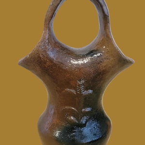 May include: A brown ceramic vessel with a glossy finish and a large circular handle at the top. The body of the container features a subtle embossed botanical design on the front, with dark, charred-looking patches near the base and handle.