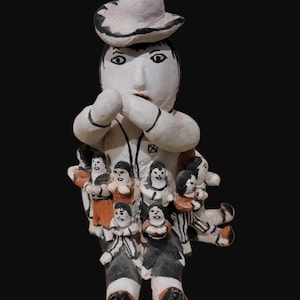 May include: A hand-painted ceramic figurine of a person wearing a hat, with a surprised expression, surrounded by several smaller figures. The figurine is primarily white with black and orange accents, set against a black background.
