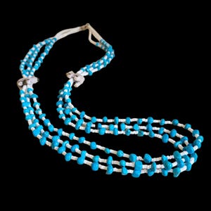 May include: A multi-strand necklace featuring turquoise-colored beads and white beads. The necklace has a layered design with three strands of beads. The beads are strung on a white cord, creating a striking contrast against the black background.