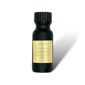 May include: A black glass bottle with a black cap, labelled "DOJO RODEO DRIVE AROMATIC DIFFUSER OIL APPROX 15 ML". The label is gold with black text. The bottle is sitting on a white surface.