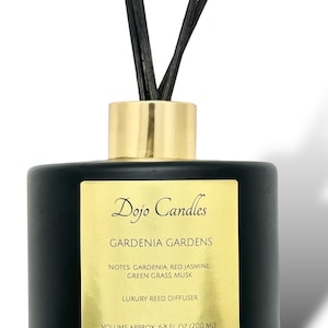 May include: Black glass reed diffuser bottle with a gold lid and black reeds. The bottle has a gold label with the text "Dojo Candles Gardenia Gardens" and the notes "Gardenia, Red Jasmine, Green Grass, Musk". The label also says "Luxury Reed Diffuser" and "Volume Approx. 6.8 fl oz (200 ml)".