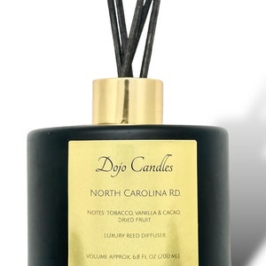 Op de afbeelding: Zwarte glazen rietdiffuserfles met een gouden label. Het label vermeldt "Dojo Candles North Carolina Rd. Notes: Tabak, Vanille & Cacao, Gedroogd Fruit Luxe Rietdiffuser Volume ca. 6,8 fl oz (200 ml)"