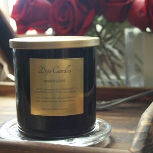 Pu&ograve; includere: Una candela in vetro nero con un coperchio e un'etichetta dorati. L'etichetta riporta "Dojo Candles" e "Marvelous" con una descrizione del profumo: "Note: Zafferano, Ribes Nero, Mora, Anice, Mirra, Incenso, Ambra. 100% Naturale, Cera di Soia Vegana. Fatto a mano in California. Peso netto 9 oz / 255 g."