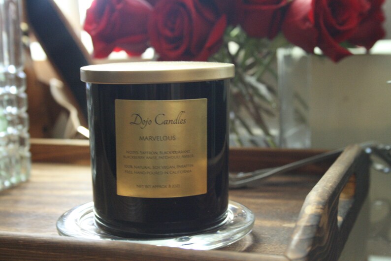 Pu&ograve; includere: Una candela in vetro nero con un coperchio e un'etichetta dorati. L'etichetta riporta "Dojo Candles" e "Marvelous" con una descrizione del profumo: "Note: Zafferano, Ribes Nero, Mora, Anice, Mirra, Incenso, Ambra. 100% Naturale, Cera di Soia Vegana. Fatto a mano in California. Peso netto 9 oz / 255 g."