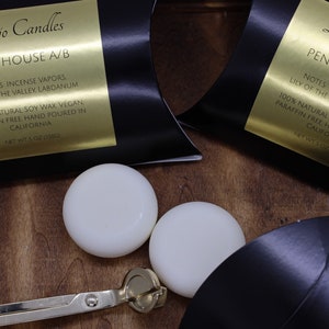 May include: Two white soy wax candles in black boxes with gold labels. The labels read "Dojo Candles Penthouse A/B" and list the notes as "Incense Vapors, Lily of the Valley, Labdanum". The candles are 100% natural soy wax, vegan, paraffin free, and hand poured in California. The boxes are on a wooden surface.