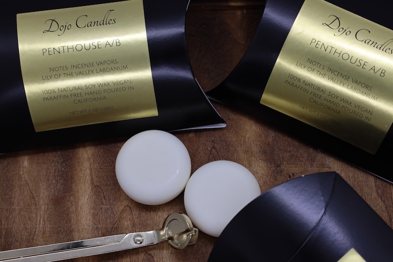 May include: Two white soy wax candles in black boxes with gold labels. The labels read "Dojo Candles Penthouse A/B" and list the notes as "Incense Vapors, Lily of the Valley, Labdanum". The candles are 100% natural soy wax, vegan, paraffin free, and hand poured in California. The boxes are on a wooden surface.