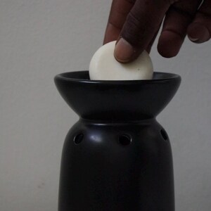May include: A black ceramic wax warmer with a white wax melt being placed inside. The warmer has a wide, round top and a narrow base.