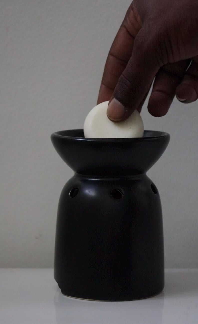 May include: A black ceramic wax warmer with a white wax melt being placed inside. The warmer has a wide, round top and a narrow base.