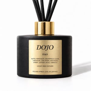 May include: A matte black reed diffuser bottle with a gold-colored collar and black reeds. The gold label features the text DOJO 1949, a list of scent notes including bergamot, grapefruit, clove, labdanum, oak wood, patchouli, amber, leather, moss, tobacco, and volume 6.8 fl oz.
