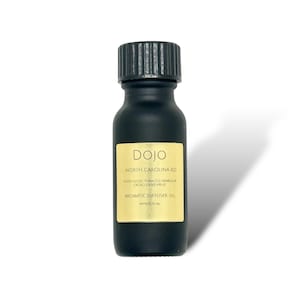 Op de afbeelding: Een zwarte glazen fles met een zwarte dop en een gouden etiket. Het etiket vermeldt "DOJO NORTH CAROLINA RD NOTES NOTES TOBACCO VANILLA & CACAO DRIED FRUIT AROMATIC DIFFUSER OIL APPROX 15 ML".