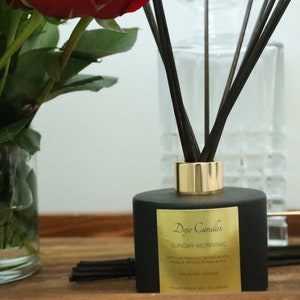 May include: Black diffuser bottle with gold lid and black reeds. The bottle has a label that reads "Dojo Candles Sunday Morning Reed & Macadamia Sandalwood, Mosco Vanilla & Winter Spices 110 ml / 3.8 oz Diffuser Volume Approx. 100 ml / 3.3 fl oz (100 ml)."