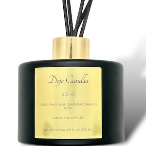 May include: Black glass bottle with a gold lid and black diffuser sticks. The bottle has a gold label with the text "Dojo Candles", "SOHO", "NOTES: WHITE ROSE, GRAPEFRUIT, OAKMOSS, MUSK", "LUXURY REED DIFFUSER", and "VOLUME APPROX. 6.8 FL OZ (200 ML)".