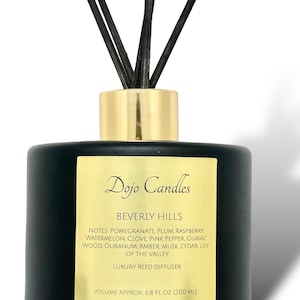 May include: Black glass reed diffuser bottle with a gold lid and black reeds. The bottle has a gold label with the text "Dojo Candles Beverly Hills" and a list of fragrance notes: Pomegranate, Plum, Raspberry, Watermelon, Clove, Pink Pepper, Guaiac Wood, Olibanum, Amber, Musk, Cedar, Lily of the Valley. The label also states "Luxury Reed Diffuser" and "Volume Approx. 6.8 fl oz (200 ml)".