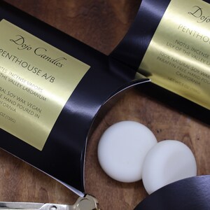 May include: Two black boxes with gold labels that read "Dojo Candles Penthouse A/B" and list the notes as "Incense Vapors, Lily of the Valley, Labdanum". The boxes are open and two white soy wax melts are visible.