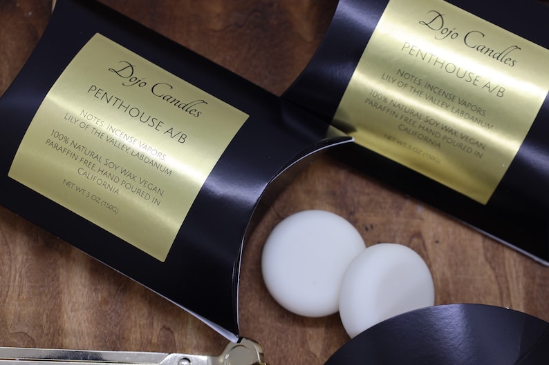 May include: Two black boxes with gold labels that read "Dojo Candles Penthouse A/B" and list the notes as "Incense Vapors, Lily of the Valley, Labdanum". The boxes are open and two white soy wax melts are visible.