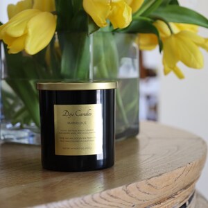 Pu&ograve; includere: Una candela nera con un coperchio e un'etichetta dorati. L'etichetta riporta "Doyo Candles" e "Marvelous". La candela &egrave; appoggiata su un tavolo di legno con un vaso di tulipani gialli sullo sfondo.
