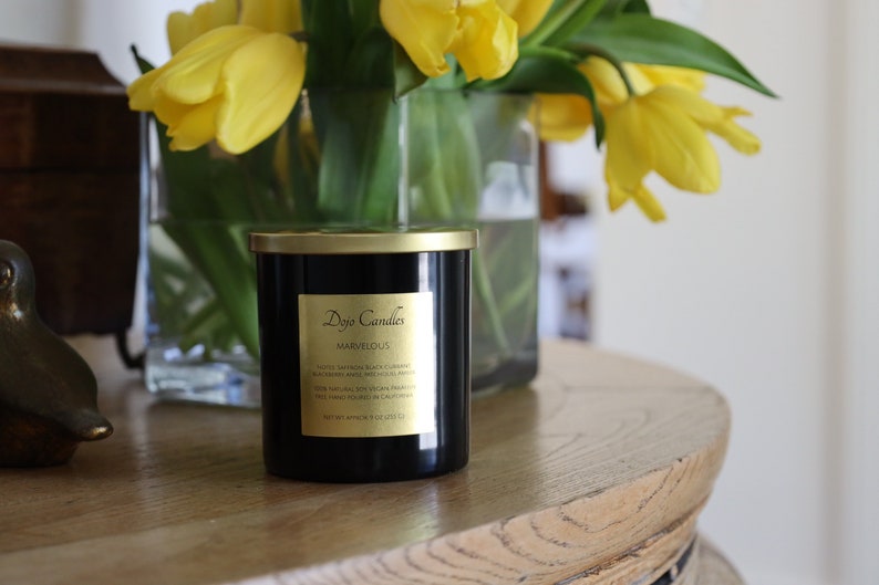 Pu&ograve; includere: Una candela nera con un coperchio e un'etichetta dorati. L'etichetta riporta "Doyo Candles" e "Marvelous". La candela &egrave; appoggiata su un tavolo di legno con un vaso di tulipani gialli sullo sfondo.