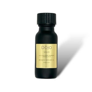 Op de afbeelding: Een zwarte glazen fles met een gouden etiket waarop "DOJO SOHO AROMATIC DIFFUSER OIL APPROX 15 ML" staat en de noten "WHITE ROSE, GRAPEFRUIT, OAKMOSS, MUSK" worden vermeld.