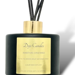 Puede incluir: Botella de difusor de juncos de vidrio negro con tapa dorada y juncos negros. La etiqueta dice "Dojo Candles Martha's Vineyard Notes: Fig Wood, Milky Sap, Green Grass Luxury Reed Diffuser Volume Approx. 6.8 fl oz (200 ml)"