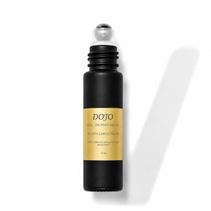 May include: A matte black glass roll-on perfume oil bottle with a gold label. The label reads DOJO, ROLL ON PERFUME OIL, NORTH CAROLINA RD, NOTES: TOBACCO, VANILLA & CACAO WOOD, FRUIT, 10 ML. The bottle features a clear plastic roller ball top.